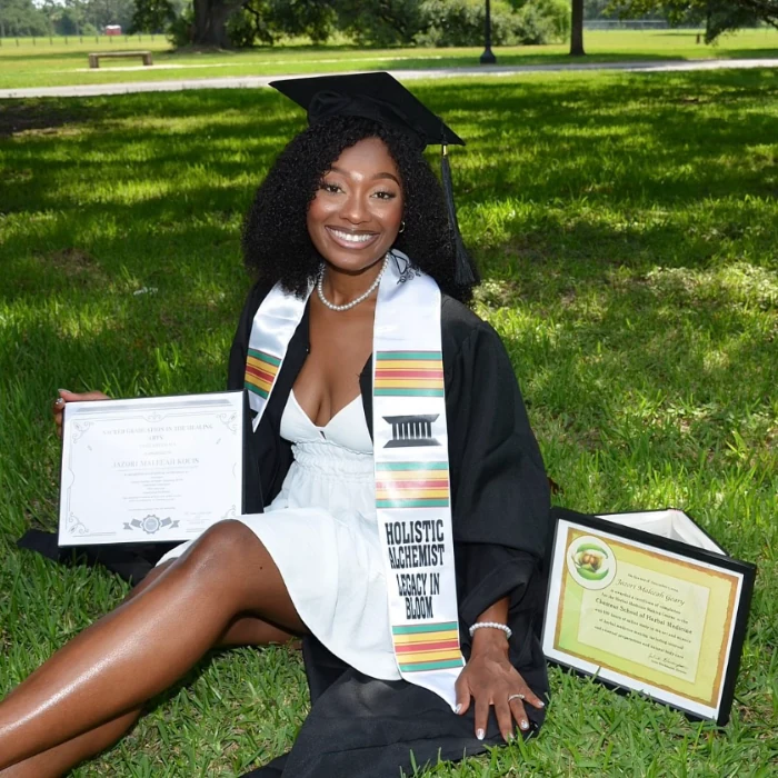 Maleeah Jay in a graduation cap and gown, smiling on the grass with two framed certificates